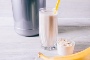 Banana, protein shake in a glass and scoop on white wooden table close-up