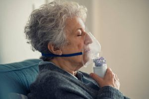 Senior woman using a nebulizer for respiratory therapy at home, eyes closed as she inhales medication.