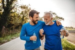 An older man and a younger man exercising outdoors, both wearing blue tops and holding water bottles. They appear to have just finished a walk and are laughing together, enjoying the moment.