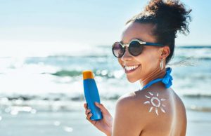 A beautiful young woman with a sun drawn on her shoulder, holding a bottle of sunscreen at the beach on a sunny day.